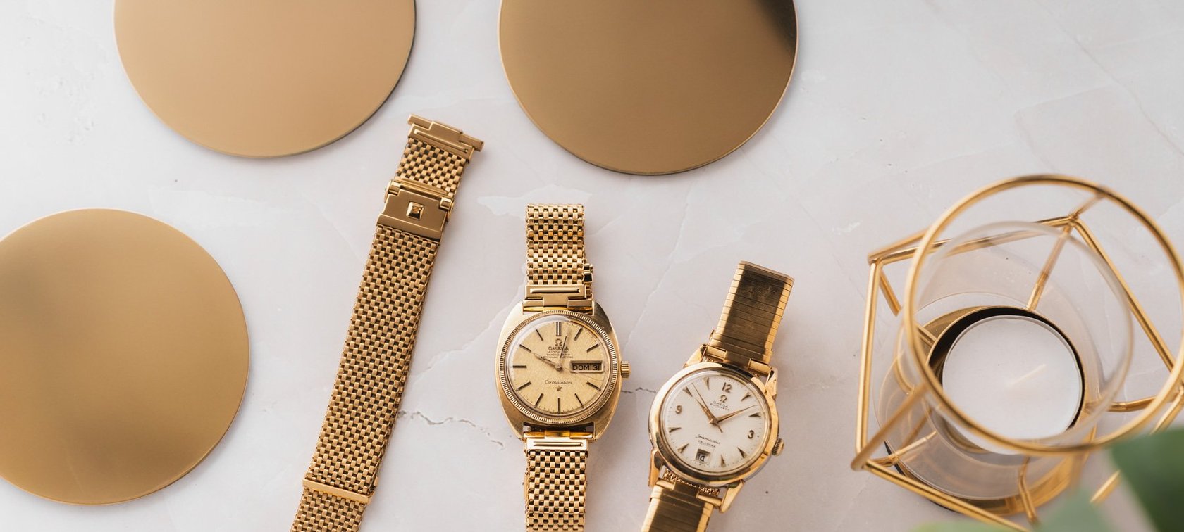 gold-plated Forstner Komfit Mesh and Military Type bracelets on Omega Constellation and Seamaster, flat-lay with gold coasters and candle