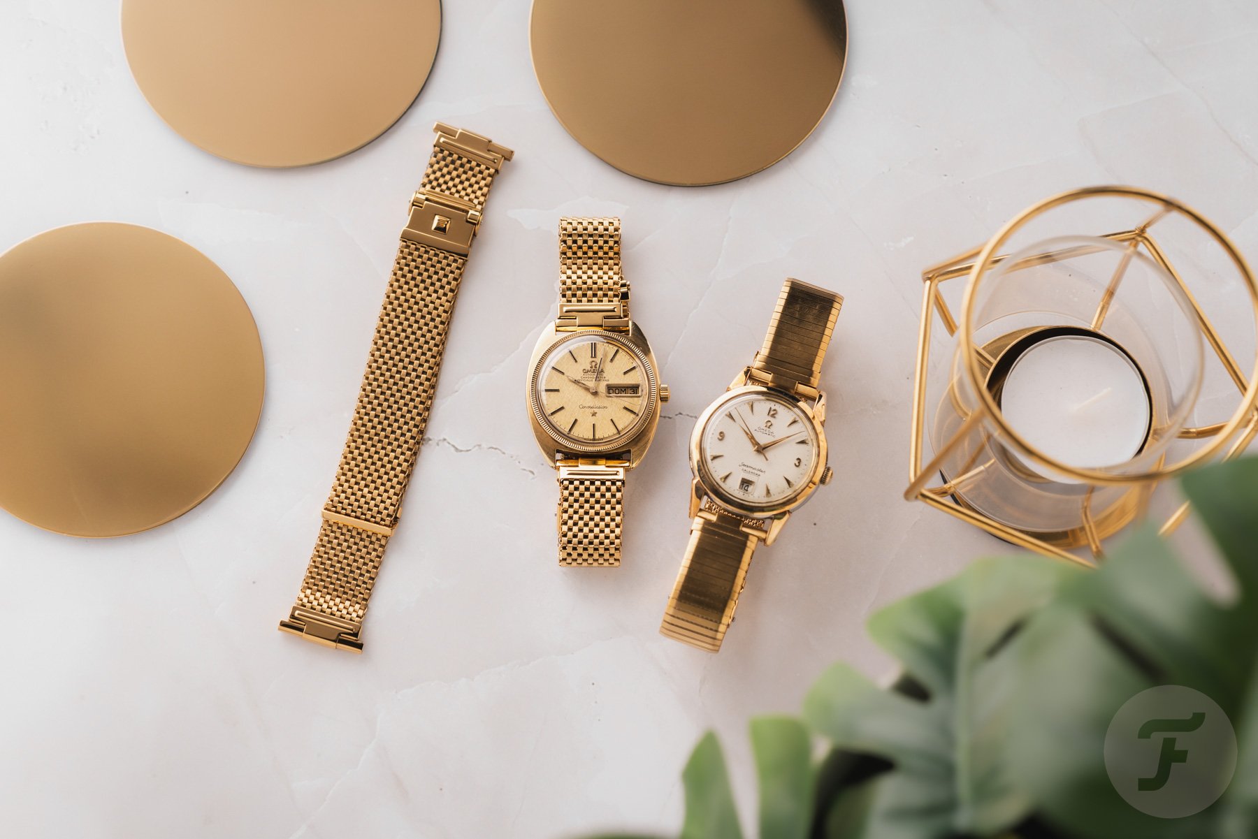 gold-plated Forstner Komfit Mesh and Military Type bracelets on Omega Constellation and Seamaster, flat-lay with gold coasters and candle
