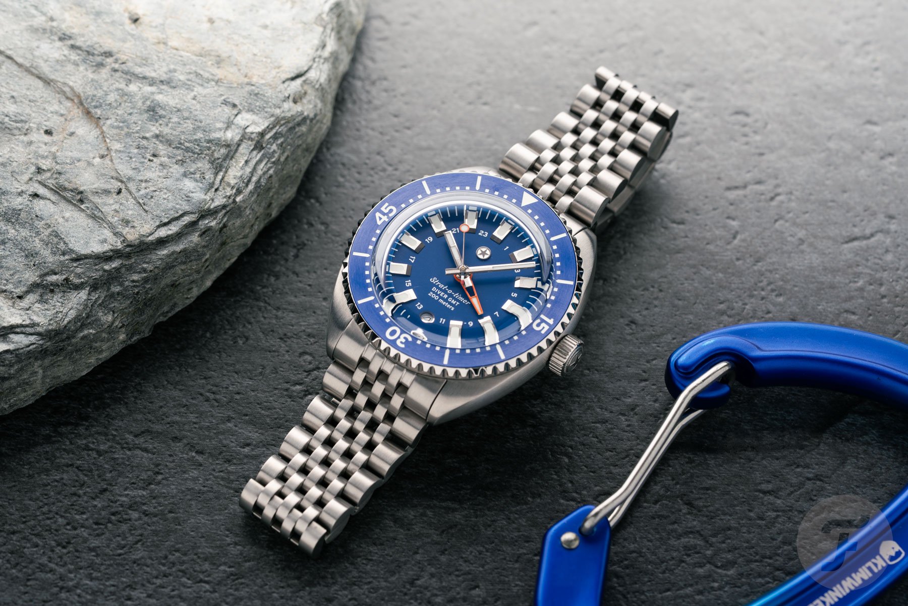 blue-dial Jack Mason Strat-o-Timer Titanium Diver GMT flat-lay between rock and blue carabiner