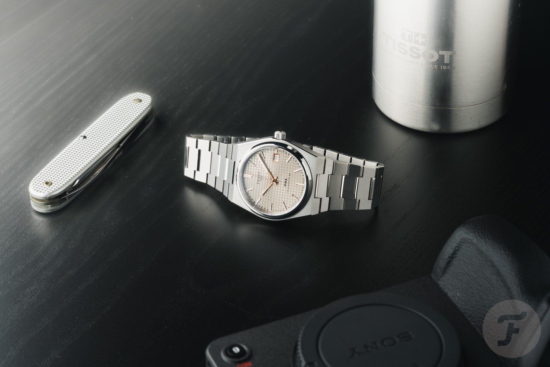 Tissot PRX Titanium 38mm on side, crown up, with pocket knife, water bottle, and camera