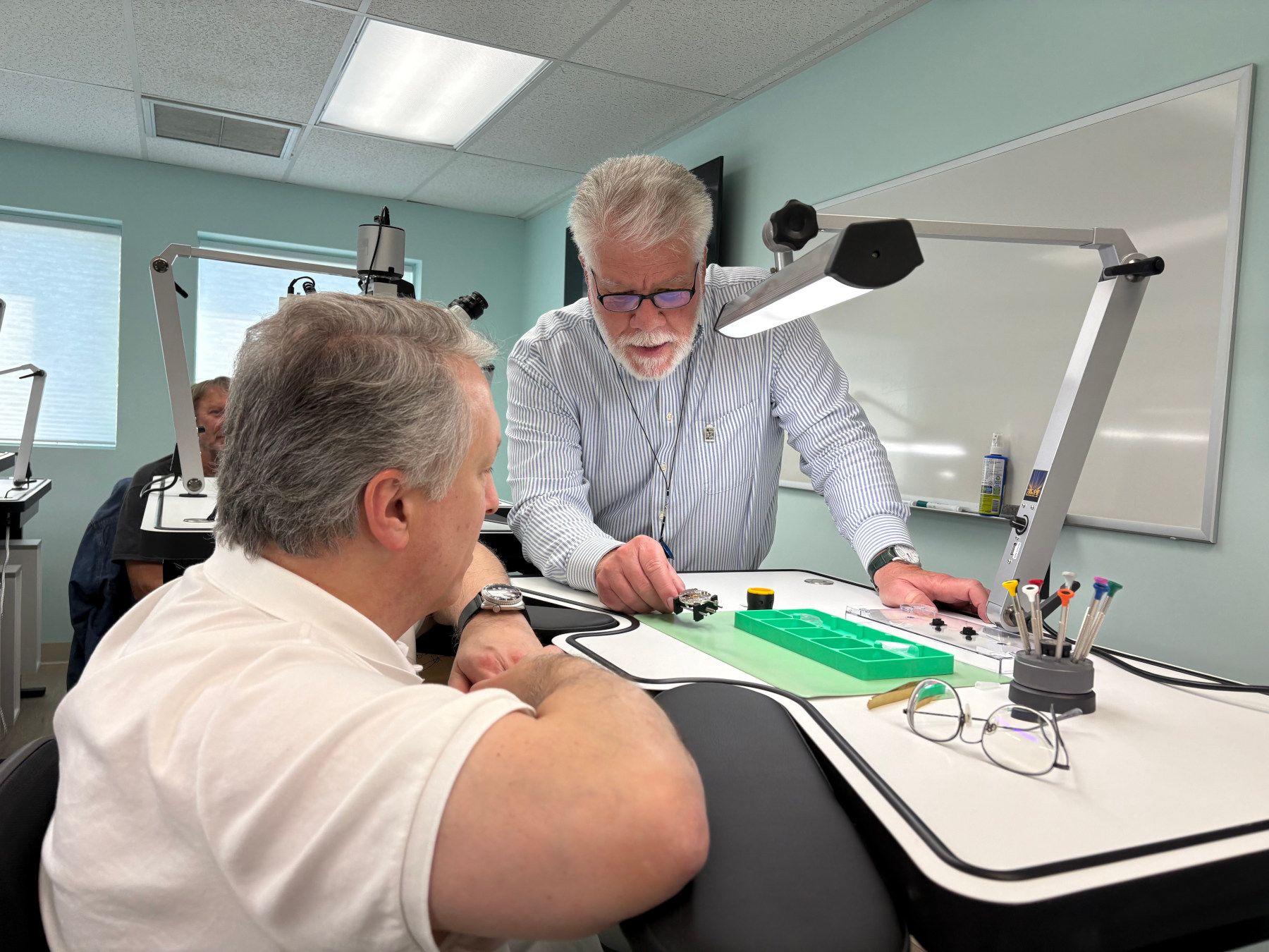 Roland Murphy speaking to a participant in a watchmaking class sat at a watchmaker's bench at Lancaster Watch Weekend